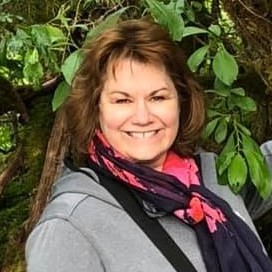 Joanne Lundberg.  Woman smiling at camera.  She has brown hair down to her shoulders, wears a multi-colored scarf and a grey coat.  She stands in front of a tree.