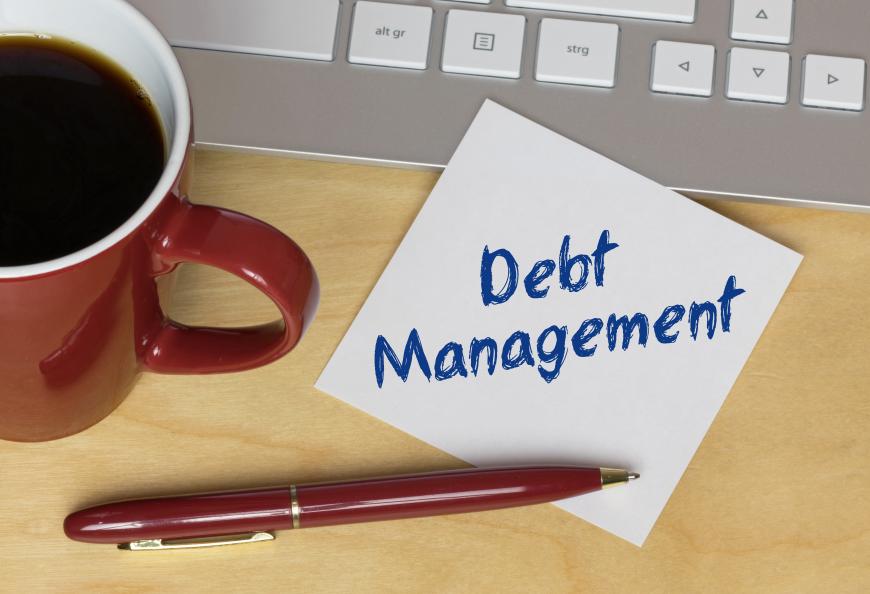 Post-It note that reads Debt Management and rests on desk. Next to note are a cup of coffee, a computer keyboard, and a red pen.