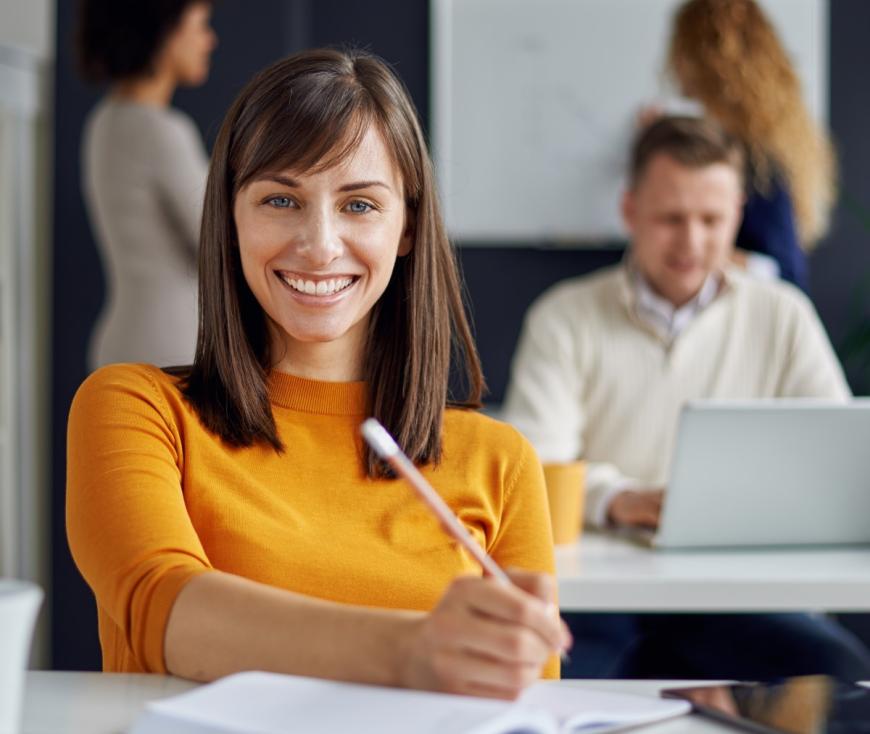 Adult woman sitting at desk, writing on papers, and smiling for the camera. Man sitting behind her and works on a laptop. Two women stand behind man in front of a whileboard.