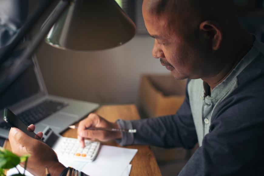 Person sits at a desk, making calculations on calculator with the right hand and holds a cell phone with the left hand. A laptop lies on the table behind the calculator.