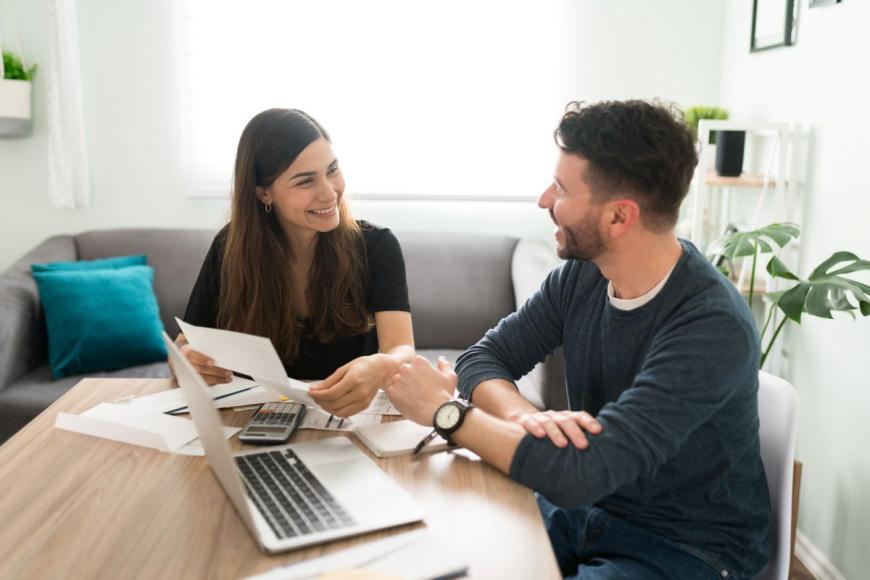 Young adult couple sitting next to each other at a table and smiling at each other. The woman is on the left and holding a piece of paper in her hand. On the table are several more papers, a laptop, a notepad, and a calculator.
