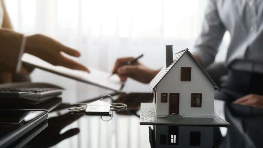 Person in a tan coat presenting a document across a table to another person who is wearing a long-sleeved, white shirt and is signing the document.  On the table are a model of a house, a set of keys, and a calculator.