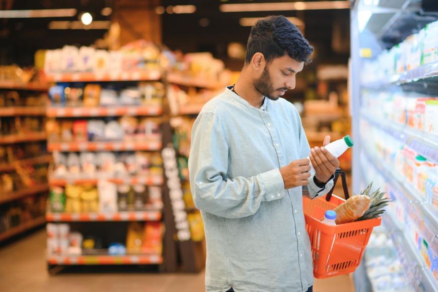 Man of South Central Asian descent in grocery store. He looks at information on a bottled item, while holds a basket with several grocery items in his left arm. He has dark, short hair and a beard, and he wears a long sleeve, light blue shirt.