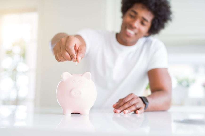 Black man smiling as he puts a coin into a light pink piggy bank. The man has an Afro hairstyle and is wearing a short-sleeved white T-shirt.