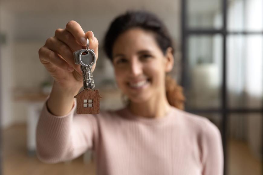 Woman holding keys to a house and smiling to the camera. She has brown hair and is wearing a pink, long sleeve top.