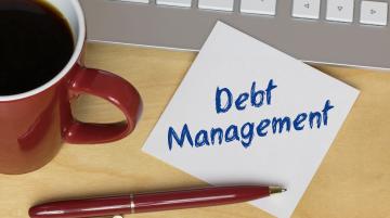 Post-It note that reads Debt Management and rests on desk.  Next to note are a cup of coffee, a computer keyboard, and a red pen.