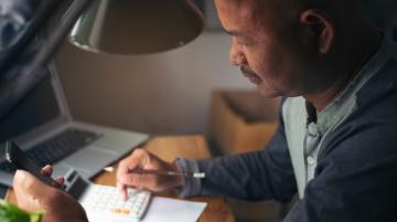 Person sits at a desk, making calculations on calculator with the right hand and holds a cell phone with the left hand.  A laptop lies on the table behind the calculator.