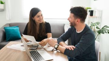 Young adult couple sitting next to each other at a table and smiling at each other.  The woman is on the left and holding a piece of paper in her hand. On the table are several more papers, a laptop, a notepad, and a calculator.