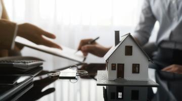 Person in a tan coat presenting a document across a table to another person who is wearing a long-sleeved, white shirt and is signing the document.  On the table are a model of a house, a set of keys, and a calculator.