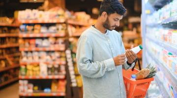 Man of South Central Asian descent in grocery store.  He looks at information on a bottled item, while holds a basket with several grocery items in his left arm.  He has dark, short hair and a beard, and he wears a long sleeve, light blue shirt.