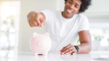 Black man smiling as he puts a coin into a light pink piggy bank.  The man has an Afro hairstyle and is wearing a short-sleeved white T-shirt.