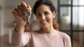 Woman holding keys to a house and smiling to the camera.  She has brown hair and is wearing a pink, long sleeve top.